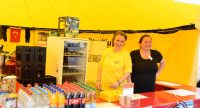 zwei Frauen in einem gelben Zelt stehen lachend vor einem roten Tisch mit Getränken drauf, Quelle: DTF