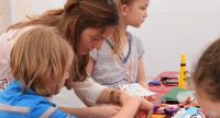 Frau steht mit Kindern an einem Basteltisch, Quelle: DTF