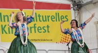 zwei Frauen in traditioneller Kleidung tanzend auf der Bühne vor Banner des Kinderfestes, Quelle: DTF