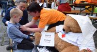 Kinder und Erwachsene sitzen zusammen neben einem Plüschbären mit Shirt des roten Kreuzes, Quelle: DTF