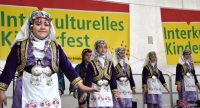 Jungs und Mädchen in traditioneller Kleidung stehen auf der Bühne vor Bannern des Kinderfestes, Quelle: DTF