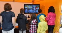 Kinder und Erwachsene in einem orangen Zelt spielen Videospiel auf einem Fernseher, Quelle: DTF