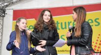 drei stehende junge Frauen auf der Bühne vor Banner des Kinderfestes sprechen ins Mikrofon, Quelle: DTF