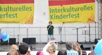 Junge in schwarzem Shirt allein auf der Bühne vor Bannern des Kinderfestes wird von Publikum beobachtet, Quelle: DTF