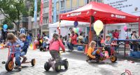 Kinder auf Holzgefährten fahren auf dem Marktplatz vor einem roten Zelt herum, Quelle: DTF