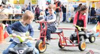 Kinder auf Holzgefährten fahren auf dem Marktplatz vor einem roten Zelt herum, Quelle: DTF