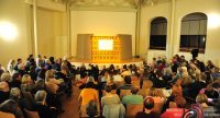 Saal voller Menschen mit hell erleuchtetem Teppich auf einem Podium in der Bildmitte, Quelle: DTF