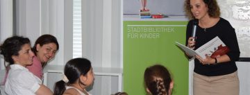 Frau steht vor sitzenden Kindern mit aufgeschlagenem Buch in den Händen