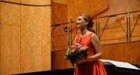 Frau in orangenem Kleid mit Blumenstrauß in den Händen lächelt verlegen Richtung Publikum, Quelle: DTF