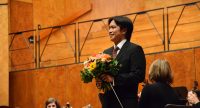 Mann im Anzug mit Blumenstrauß in den Händen lächelt Richtung Publikum, Quelle: DTF