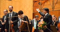Orchester auf dcer Bühne, der Dirigent erhebt den rechten Arm als Gruß zum Publikum hoch, Quelle: DTF