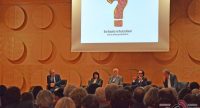Bühne mit sitzenden Diskussionsteilnehmern und Leinwand mit projiziertem Fragezeichen, Publikum im Bildvordergrund, Quelle: DTF