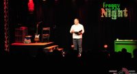 Mann im weißen Hemd hält sprechend einen Ordner in seinen verschränkten Händen auf Bühne vor Silhouette des Publikums, Quelle: DTF