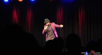 Mann im pinken Sakko mit schwarzem Hut spricht ins Mikrofon vor Silhouette des Publikums, Quelle: DTF
