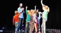 Kinder auf der Bühne erheben die Arme, Mann im blauen Hemd steht im Bildhintergrund, Quelle: DTF
