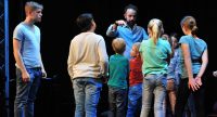 Kinder auf der Bühne erheben die Arme, Mann im blauen Hemd steht im Bildhintergrund, Quelle: DTF