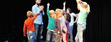 Kinder auf der Bühne erheben die Arme, Mann im blauen Hemd steht im Bildhintergrund