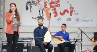 zwei sitzende Musiker auf der Bühne vor Banner des Kinderfestes, neben ihnen spricht eine Frau im orangen Sakko ins Mikrofon, Quelle: DTF
