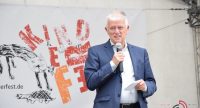 Mann im blauen Sakko spricht ins Mikrofon auf Bühne vor Banner des Kinderfests, Quelle: DTF