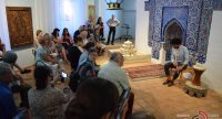 sitzende Menschen schauen einem Mann mit traditionellem Saiteninstrument vor blauer Arabeske zu, Quelle: DTF