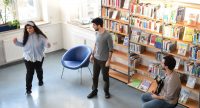 zwei junge Männer und ein Mädchen vor einem Bücherregal, Quelle: DTF