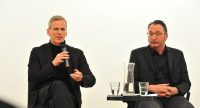 zwei Herren in schwarzen Anzügen sitzend an einem Tisch schauen nach vorne ins Publikum sprechend, Quelle: DTF