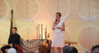 Frau in weißem Kleid spricht ins Mikrofon auf der Bühne vor Köpfen des Publikums, Quelle: DTF