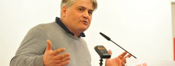 Mann im grauen Pullover spricht gestikulierend ins Mikrofon an einem Rednerpult