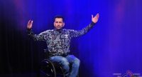 Mann im Rollstuhl und Tarnfarben Oberteil hebt die Arme in die Luft, Quelle: DTF