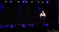Frau in schwarzem SHirt mit pinker Jacke zeigt mit den Händen nach unten vor Silhoeutte des Publikums, Quelle: DTF