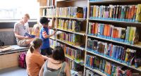 Kinder und Frauen stehen vor einem Regal voller bunter Bücher, Quelle: DTF