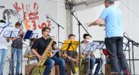 Saxophonspielende Kinder auf der Bühne sitzend mit älterem Dirigenten im blauen Shirt, Quelle: DTF