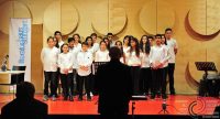 Kinderchor in weißen Hemden auf Bühne vor Silhouette des Chorführers, Quelle: DTF
