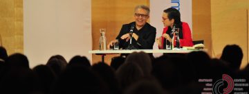 Mann und Frau sitzend auf der Bühne vor Banner des DTF und des Literaturhauses und SIlhouette des Publikums, der Mann lacht