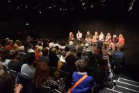 Frauen im Kostüm auf der Bühne sitzend auf Stühlen vor Publikum, Quelle: DTF