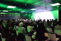 Saal voller Menschen auf weißen Stühlen vor der stark grün beleuchteten Bühne, Quelle: DTF