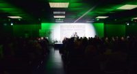 Menschen im dunklen Saal schauen Autor auf grün bleuchteter Bühne zu, Quelle: DTF