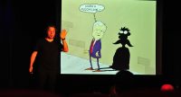Mann im schwarzen T-Shirt auf der Bühne hält die linke Hand ans Ohr vor einer Karikatur, welche einen Mann im ANzug mitsamt Schatten zeigt, Quelle: DTF