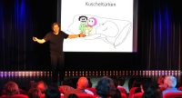 Mann im schwarzen T-Shirt auf der Bühne hält Arme weit von sich gestreckt neben der weißen Leinwand mit einer Karikatur mit dem Titel Kuscheltürken, Quelle: DTF