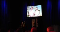 Bildschirm zweier Männer in weißen Shirts vor Silhouette des rot beleuchteten Publikums, Quelle: DTF