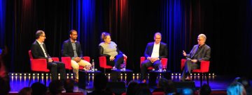 Fünf Diskussionteilnehmer sitzend auf roten Stühlen vor schwarzem Vorhand und Silhouette des Publikums im Bildvordergrund