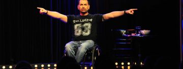 Mann im schwarzen T-Shirt auf einem Rollstuhl zeigt mit den Händen zur Seite