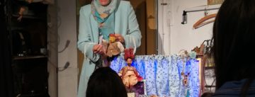 Frau im türkisen Mantel mit Kopftuch steht am Puppentheater