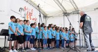Kinderchor in blauen T-Shirts mit Chorleiter in grauem sportlichem T-Shirt auf der weißen Bühne, Quelle: DTF