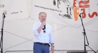 Mann in weißem Hemd spricht ins Mikrofon auf der Bühne mit dem Banner des Kinderfestes im Hintergrund, Quelle: DTF