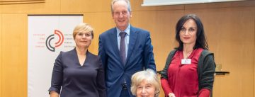 DTF Vorstandsvorsitzende, Mann im blauen Sakko und Frau im roten Oberteil verleihen Preis an sitzende ältere Frau