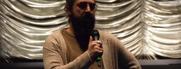 Schauspieler spricht ins Mikrofon, er hat die linke Hand in die Hüfte gestemmt