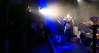 Musiker auf hell beleuchteter Bühne vor Masken-tragenden Zuschauer*innen, Quelle: DTF