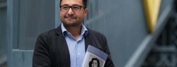 Abdul Vahap Cilhüseyin  mit einem Buch in den Händen vor einem blau-grauen Zaun