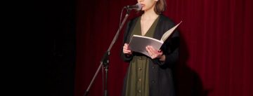 Maria Tramountani liest aus einem Buch vor, sie steht auf der Bühne vor einem roten Vorhang